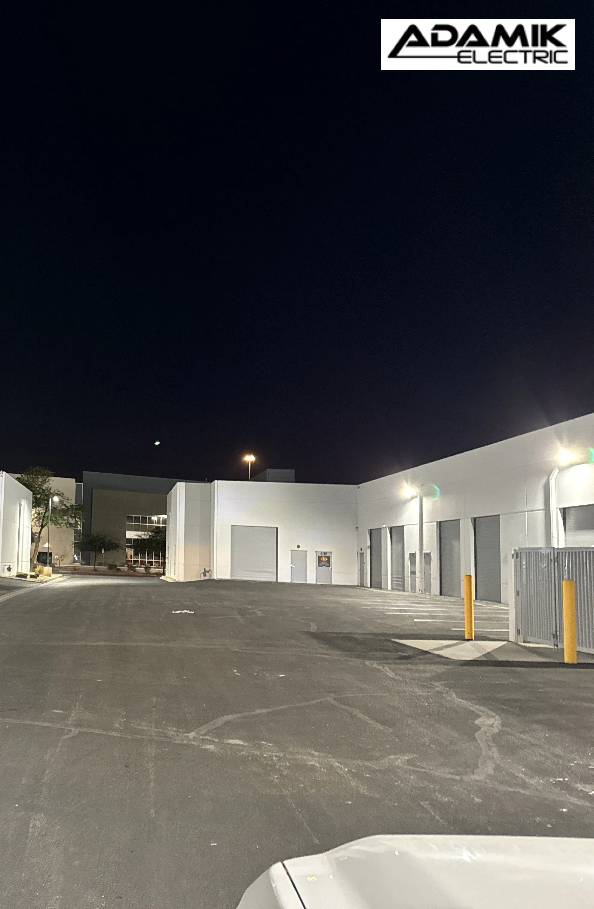 A nighttime view of a commercial facility in North Las Vegas with multiple white buildings and closed garage doors. The logo "ADAMIK ELECTRIC" is visible in the top right corner, highlighting the company’s expert electric service in the area.
