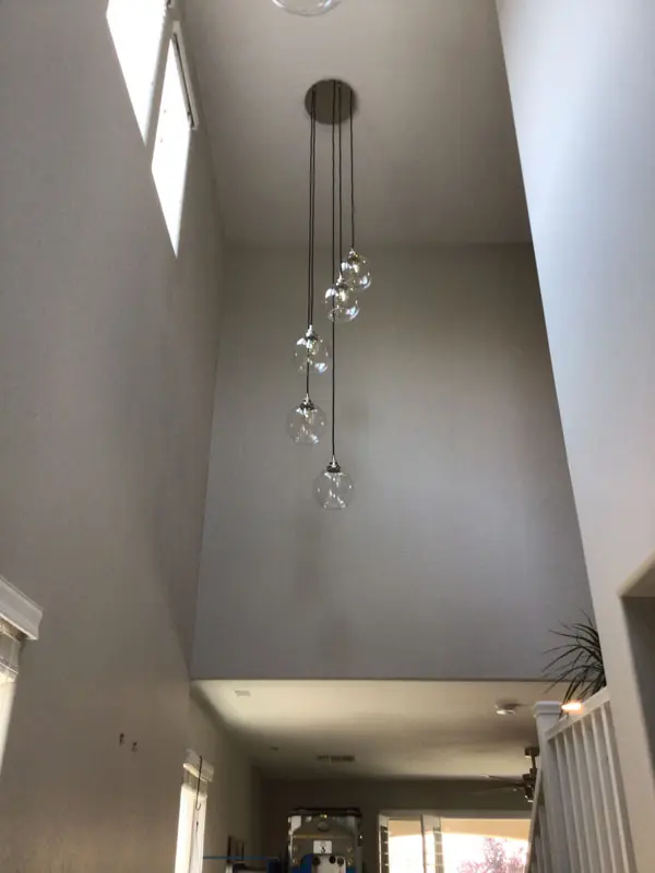A modern entryway with high ceilings features a contemporary chandelier hanging with multiple glass orbs at varying heights, installed flawlessly by Adamik Electric in North Las Vegas. Natural light streams through the windows.