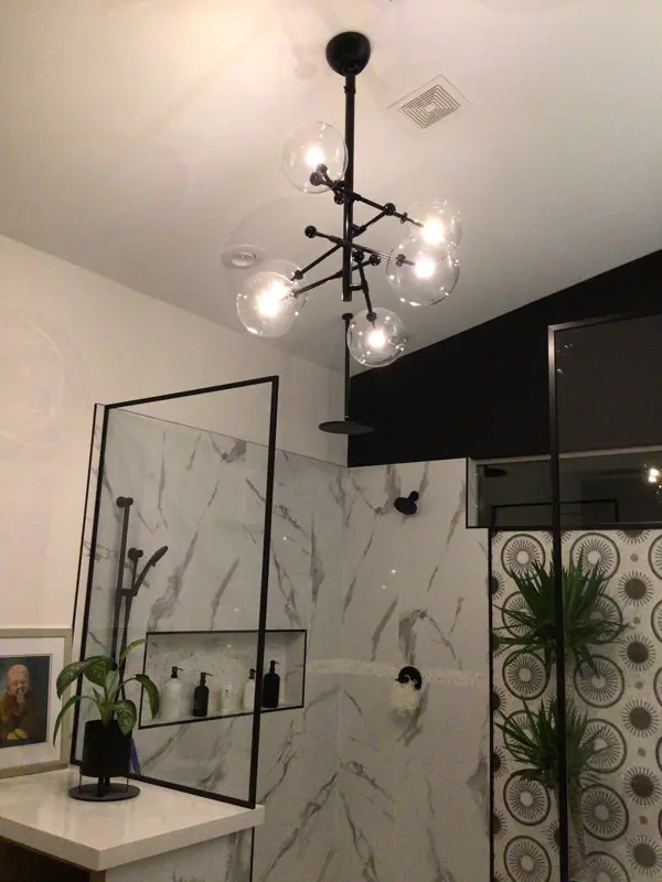 A modern bathroom in North Las Vegas features a marble-patterned shower with a glass door, a black and white geometric wall design, a potted plant on the counter, and a chandelier with multiple round light bulbs installed by Adamik Electric.