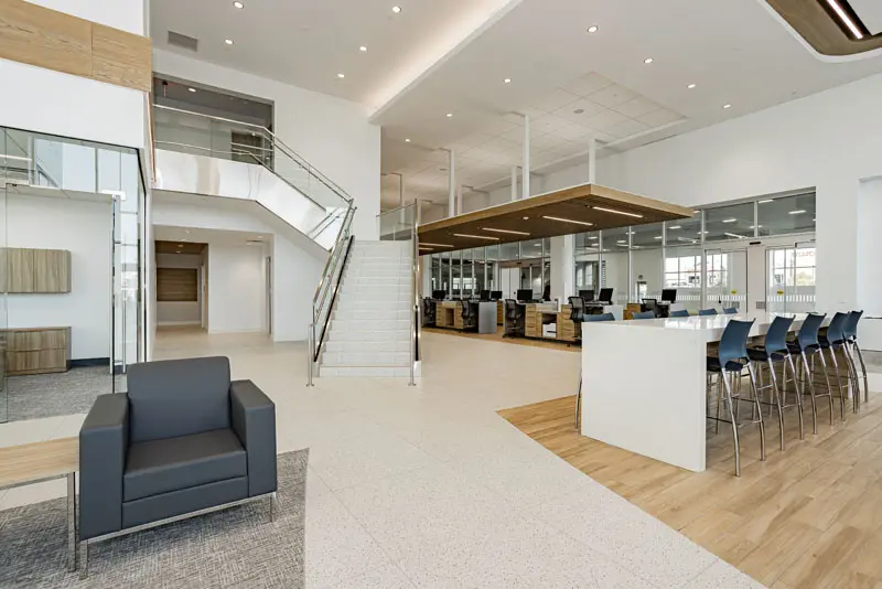 Spacious modern office space in North Las Vegas with an open floor plan, featuring a stairway, rows of workstations, a lounge area with chairs, and a bar table with high stools. Bright lighting and large windows. Conveniently equipped with Tesla Charging stations onsite for electric service.