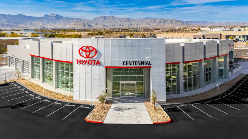 An exterior view of a modern Toyota dealership named Centennial in North Las Vegas, with mountains in the background and an empty parking lot in the foreground.