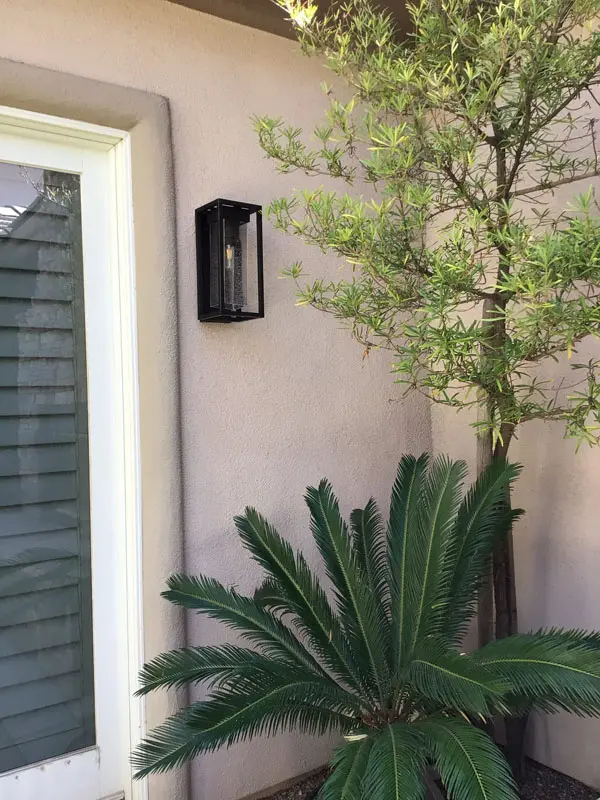 A wall-mounted lantern light next to a glass door illuminates a small green tree and palm plant in a garden, enhancing the serene ambiance of this North Las Vegas property.