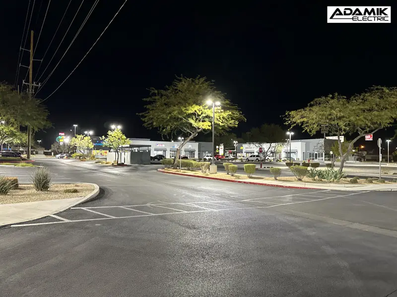 Nighttime view of a well-lit, empty parking lot with trees, a few parked cars, and surrounding businesses illuminated in the background, thanks to the reliable work of Adamik Electric providing top-notch electric service in North Las Vegas.
