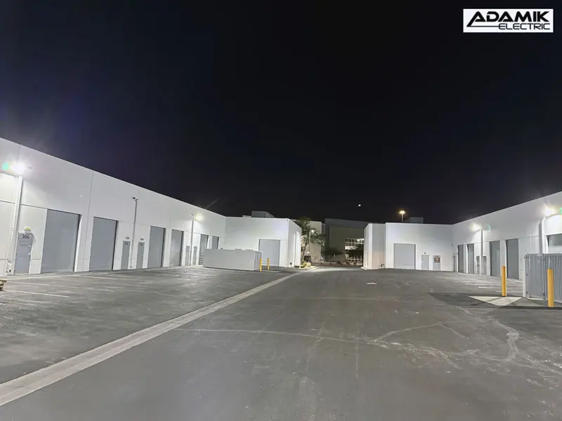 Night view of an empty commercial complex in North Las Vegas with multiple closed garages, illuminated by exterior lights. The Adamik Electric logo is in the top right corner, proudly showcasing their top-notch electric service.