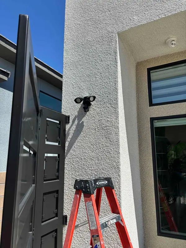 A red ladder, courtesy of Adamik Electric, is positioned under a mounted security camera with two floodlights on the exterior wall of a building near a window and an open gate in North Las Vegas.