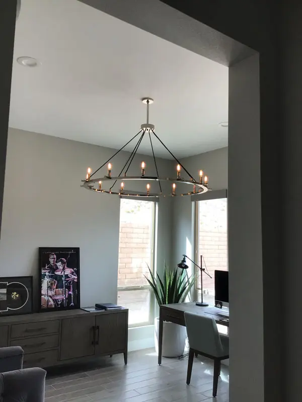 A modern living room in North Las Vegas with a large circular chandelier, a wooden desk and chair, a plant, and natural light from large windows. A framed picture and decor items are on a console table against the wall. Design by Adamik Electric ensuring excellent electric service throughout.